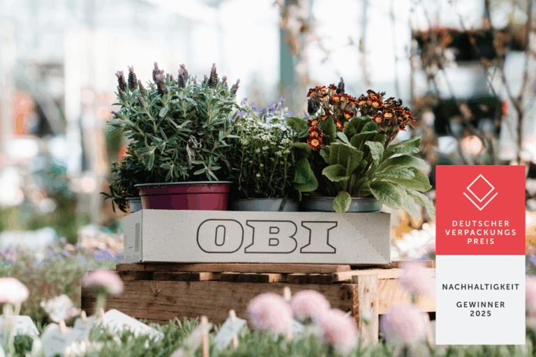Das Bild zeigt ein leerer grauer Karton mit dem Logo "OBI" auf der Vorderseite. Der Karton steht auf einer Holzpalette, die aus mehreren Brettern zusammengesetzt ist. Im Hintergrund sind zahlreiche bunte Blumen und Pflanzen zu sehen, die eine lebendige und fröhliche Atmosphäre schaffen. Die Farben im Hintergrund sind überwiegend gelb und lila, was die Kiste im Vordergrund hervorhebt. Das Licht ist warm und freundlich, was die gesamte Szene einladend wirken lässt.
