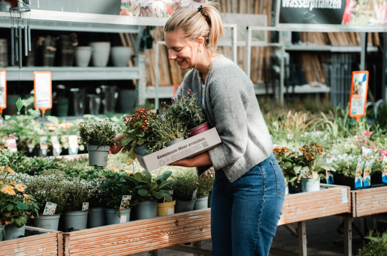Das Bild zeigt eine Person, die in einem Gartencenter steht und einen Karton mit Pflanzen trägt, den sie gerade befüllt. Auf dem Karton steht "Gemeinsam Moor erreichen". Die Person hat lange, blonde Haare, die zu einem Pferdeschwanz gebunden sind, und trägt einen grauen Pullover sowie Jeans. In der Kiste sind mehrere Töpfe mit verschiedenen Pflanzen, darunter bunte Blumen, sichtbar. Im Hintergrund sind viele weitere Pflanzen und Töpfe zu sehen, die eine grüne und blühende Umgebung schaffen. Es gibt auch Preisschilder und Beschilderungen, die auf die angebotenen Produkte hinweisen. Das Licht ist hell und freundlich, was die Szene einladend macht.