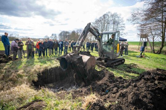 Eine Gruppe von Menschen steht auf einer Wiese um einen Bagger, der ein Loch im Boden gräbt – symbolischer Spatenstich zur Wiedervernässung eines Moores (Sernitz) bei Angermünde.